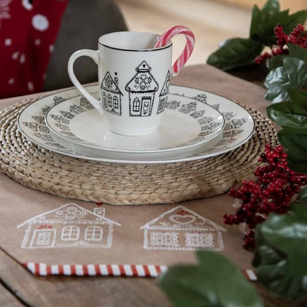 Ein weihnachtlich gedeckter Tisch mit einer Stoffserviette „Lebkuchenhäuser“ von Clayre &amp; Eef, einer Tasse und Untertasse mit Lebkuchenhausmotiven, einem Pfefferminz-Zuckerrohr in der Tasse und festlicher Dekoration drumherum.