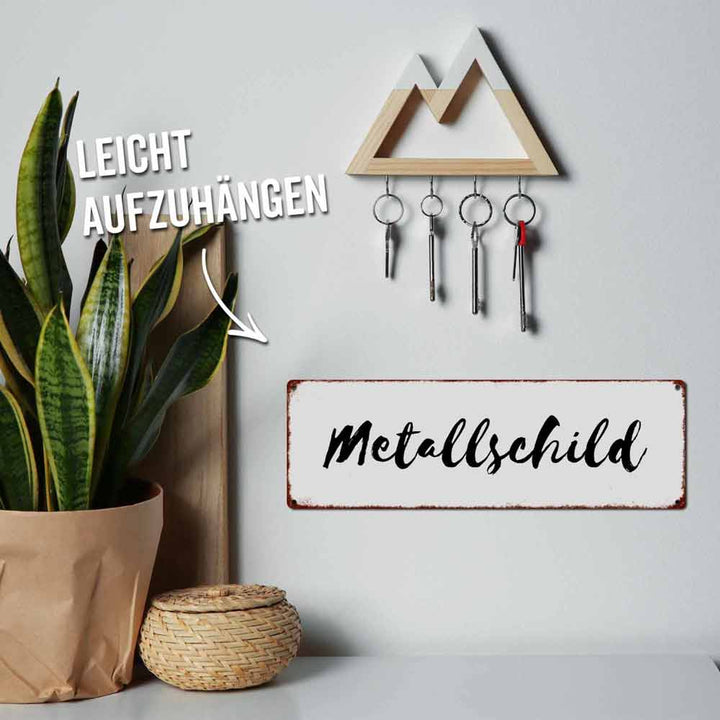 Ein an der Wand befestigtes Schlüsselbrett mit vier hängenden Schlüsseln, einer Topfpflanze und einem Metallschild mit der Aufschrift „Interluxe Metallschild – Mama, warum putzt du?“. Die Aufschrift „Leicht aufzuhängen“ weist auf das Schlüsselbrett hin.