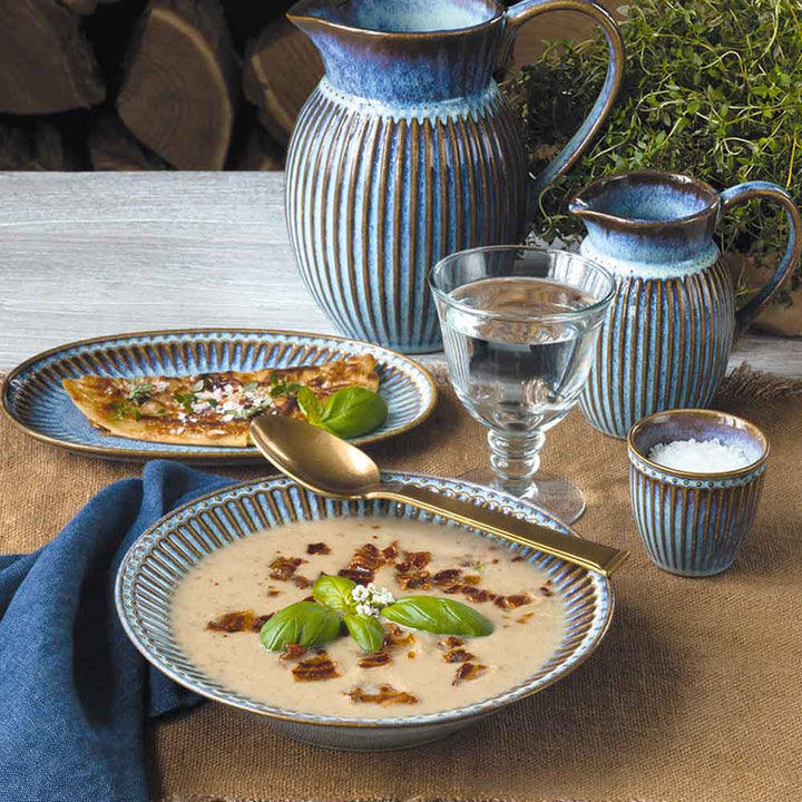Ein rustikaler Tisch mit einer Schüssel Cremesuppe, garniert mit Kräutern, begleitet von GreenGate - Alice Eierbecher-Geschirr, einem Glas Wasser und einer blauen Textilserviette.